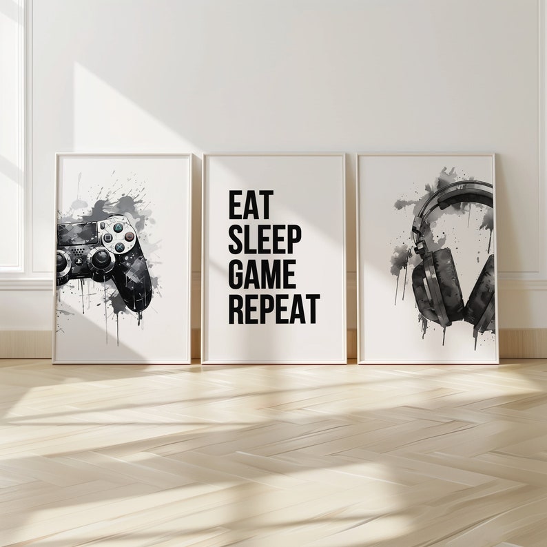 May include: Three black and white prints with a gamer theme. The first print features a black video game controller with white splatters of paint. The second print has the text "Eat Sleep Game Repeat" in black. The third print features a pair of black headphones with white splatters of paint.