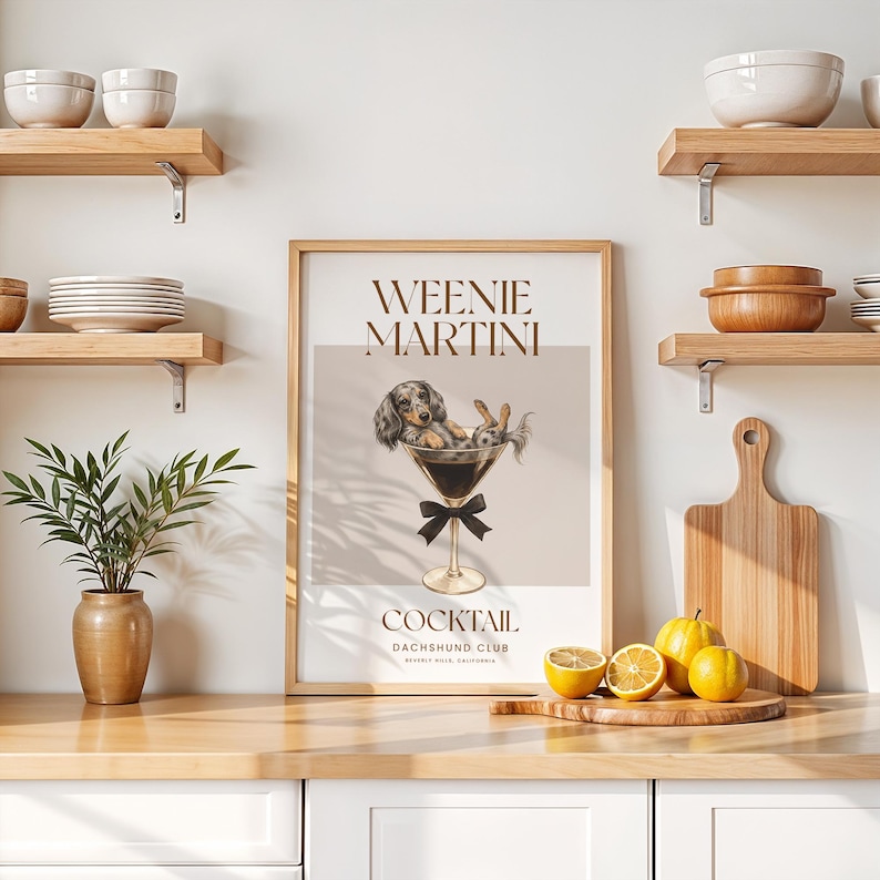 May include: A framed print featuring a dachshund dog and a rabbit in a martini glass, with the words "Weenie Martini" and "Cocktail" in brown text. The print is displayed on a wooden countertop with lemons and a cutting board.