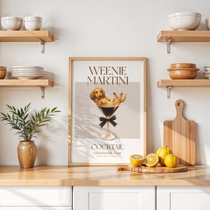 May include: A framed print featuring a dachshund dog relaxing in a martini glass, with the words "Weenie Martini Cocktail" above and below the image. The print has a light brown frame and is displayed on a wooden surface.