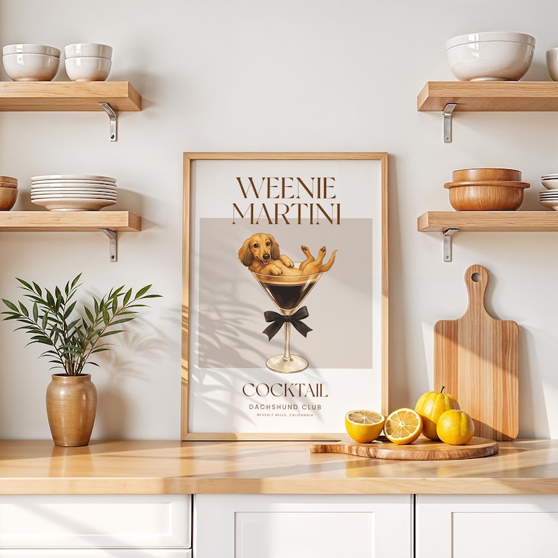 May include: A framed print featuring a dachshund dog relaxing in a martini glass, with the words "Weenie Martini Cocktail" above and below the image. The print has a light brown frame and is displayed on a wooden surface.