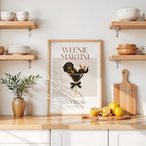 May include: A framed art print featuring a dachshund dog relaxing in a martini glass. The print has the words "Weenie Martini" and "Cocktail Dachshund Club" in a stylish font. The print is displayed on a wooden countertop with lemons and a cutting board.