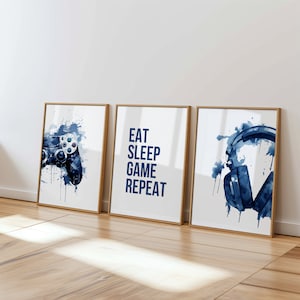 May include: Three framed prints with blue watercolour designs. The left print features a video game controller, the middle print has the text "Eat Sleep Game Repeat", and the right print shows a pair of headphones.