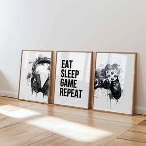 May include: Three framed prints with a black and white watercolor design. The left print features a pair of headphones, the middle print has the text "Eat Sleep Game Repeat", and the right print shows a video game controller.