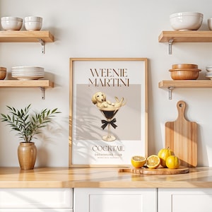 May include: A framed print with the words "Weenie Martini" and a cartoon dachshund dog relaxing in a martini glass. The print has a light brown background and a black bow tie. The print is on a wooden surface with lemons and a cutting board.