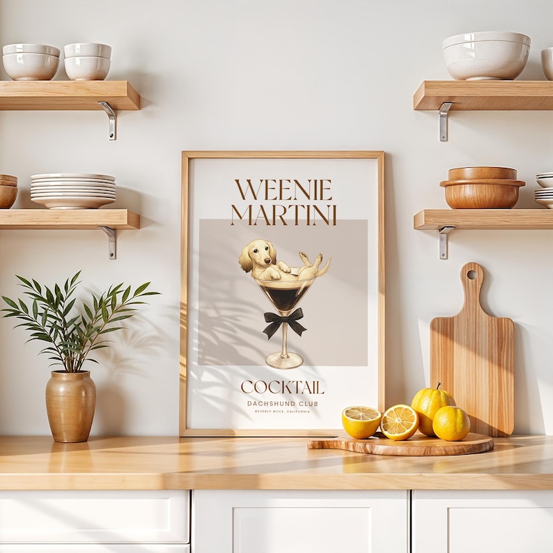 May include: A framed print with the words "Weenie Martini" and a cartoon dachshund dog relaxing in a martini glass. The print has a light brown background and a black bow tie. The print is on a wooden surface with lemons and a cutting board.