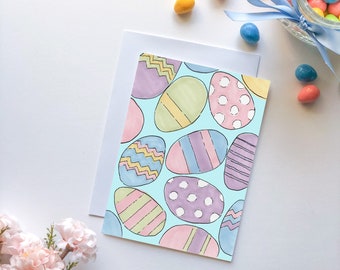 May include: A blank white card with a blue background and colorful Easter eggs. The eggs are decorated with stripes, polka dots, and zigzags. The card is surrounded by pastel-colored candy.