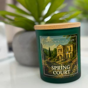 May include: A green glass candle jar with a wooden lid. The label reads "THE COURTS COLLECTION SPRING COURT" and features a painting of a building and garden. A green plant is in the background.