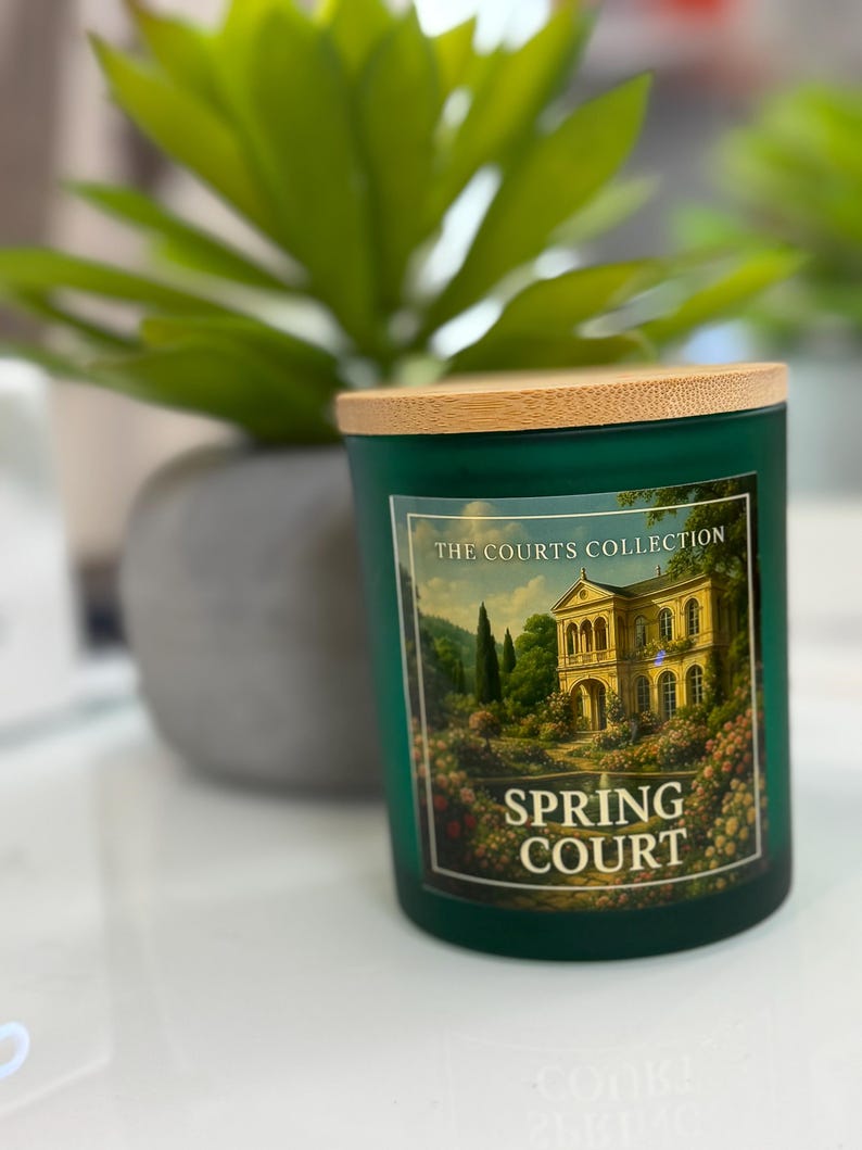 May include: A green glass candle jar with a wooden lid. The label reads "THE COURTS COLLECTION SPRING COURT" and features a painting of a building and garden. A green plant is in the background.