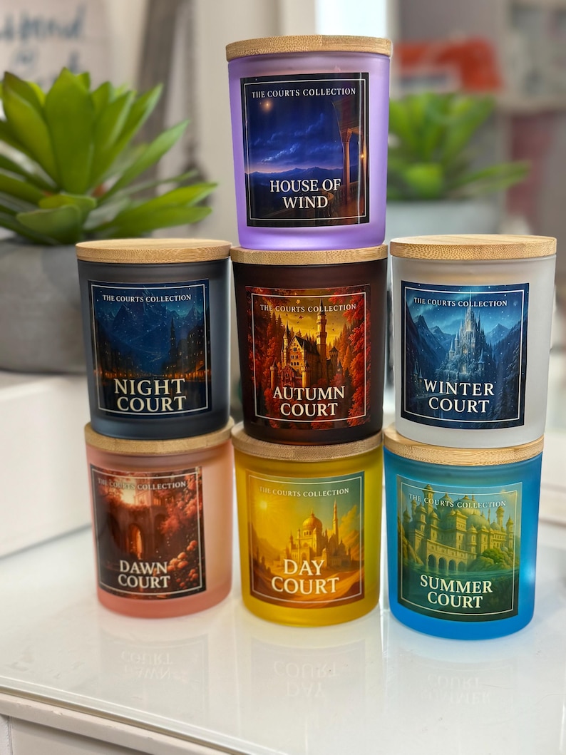 May include: A stack of seven glass candles with wooden lids. Each candle has a different color and label with the text "The Courts Collection" and a court name: House of Wind, Night Court, Autumn Court, Winter Court, Dawn Court, Day Court, and Summer Court.