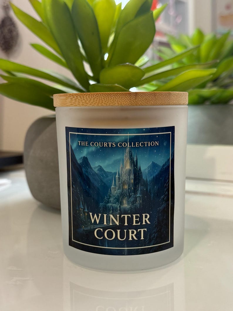 May include: A frosted glass candle with a wooden lid. The label reads "THE COURTS COLLECTION" and "WINTER COURT" with a scenic illustration of a castle in a mountain landscape. The candle is placed in front of a green plant.