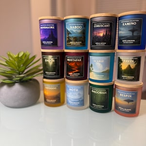 May include: A collection of soy wax candles in glass jars with wooden lids. Each candle features a different Star Wars planet name, such as "Mandalore" and "Naboo", with corresponding artwork. The candles are arranged on a white surface.