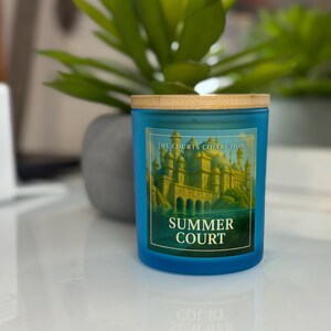 May include: A blue candle with a wooden lid. The label reads "The Courts Collection" and "Summer Court" with an illustration of a castle. The candle sits on a white surface with a green plant in the background.