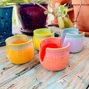 May include: A collection of handmade ceramic sponge holders in various colors, including orange, yellow, and purple. Each holder has a unique curved design and a glazed finish, sitting on a weathered wooden surface. The holders are designed to hold a sponge.