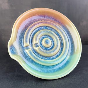 May include: A round ceramic spoon rest featuring a concentric circular maze pattern. The glaze transitions from a warm amber and tan at the outer rim to deep shades of blue and teal in the centre, with a small spout on the left side of the dish.