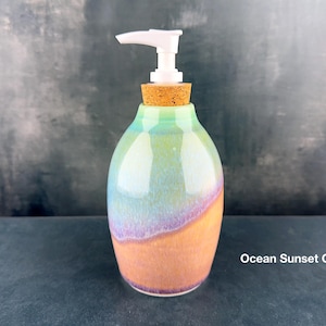 May include: A ceramic soap dispenser with an ocean sunset glaze. The dispenser has a white pump, a cork stopper, and a colorful gradient of green, purple, and orange. The text "Ocean Sunset Glaze" is visible.