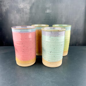 May include: Four ceramic mugs with a layered glaze design. The mugs are in shades of pink, green, yellow, and blue.