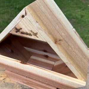 May include: A wooden birdhouse with a triangular roof and an open front. The birdhouse is made of light-colored wood and features the word "airbnb" with bird illustrations. The birdhouse is sitting on a wooden surface.
