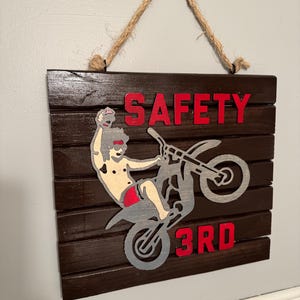Puede incluir: Un letrero de madera con fondo marrón y una silueta gris de una persona montando una moto de cross. El letrero dice "SAFETY 3RD" en letras rojas.