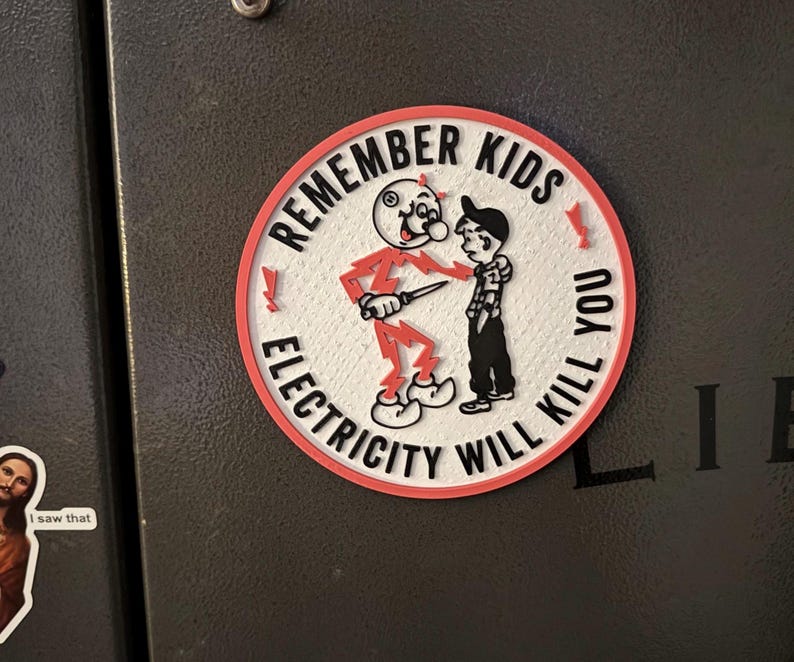 May include: A red and white round sticker with the text "Remember Kids Electricity Will Kill You". A cartoon figure of a red devil holding a knife is shown next to a boy.