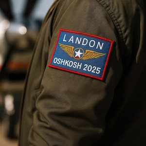 May include: An olive green bomber jacket sleeve with a rectangular patch. The patch, with a blue background and gold wings, reads "LANDON" and "OSHKOSH 2025". The jacket is a classic style, suitable for various occasions.