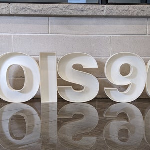 May include: White, freestanding letters spelling "LOIS 90" on a tiled floor. The letters are hollow and have a 3D effect.