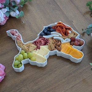 Peut inclure: Un plateau de service en céramique blanche en forme de lion avec des compartiments remplis de diverses collations, notamment des craquelins, du fromage, des raisins, du salami et des amandes.