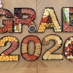 Peut inclure: Un plateau de charcuterie formant "GRAD 2026". Les lettres et chiffres sont remplis de viandes, fromages, crackers, olives, raisins et autres collations. L'arrangement est sur une surface en bois, avec des décorations florales.
