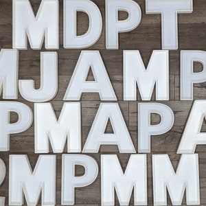 May include: White letters of the alphabet, including M, D, P, T, J, A, and others, are arranged in a random pattern on a wood background.