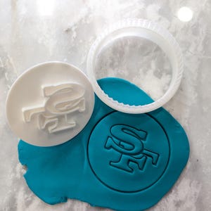 May include: A white cookie cutter and stamp with the letters "SF" are next to a teal-colored circle of playdough with the same imprint. The cookie cutter is round with a ridged edge.