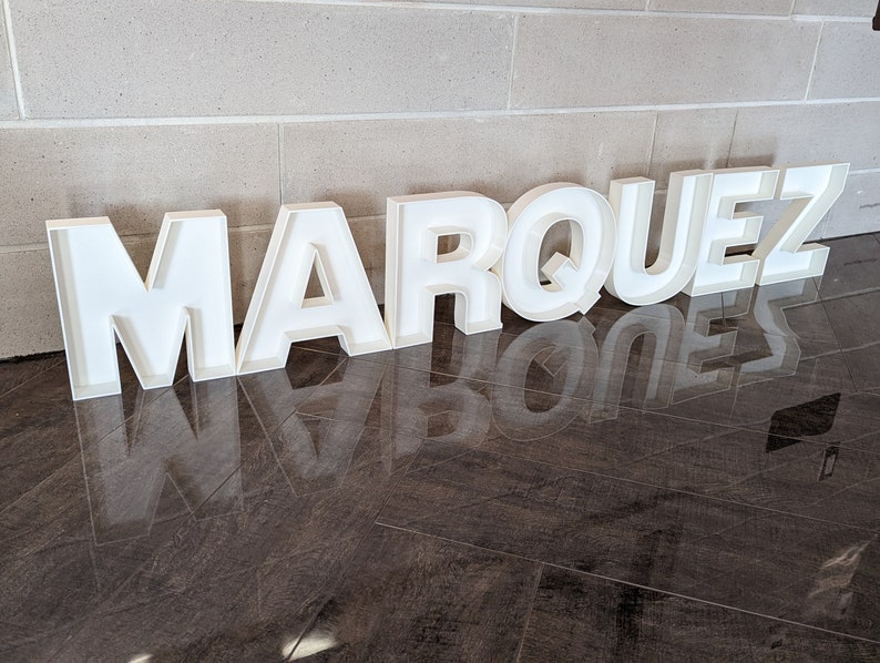 May include: White, freestanding letters spelling out the name "MARQUEZ" on a dark brown tiled floor.