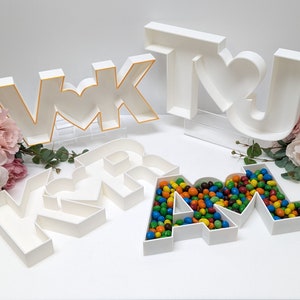 May include: White plastic letters spelling "VK" and "AM" with a heart shape in the middle of each letter. The "VK" letters are on a clear acrylic stand. The "AM" letters are filled with colorful candy. The "U" letter is also white plastic and has a heart shape in the middle.