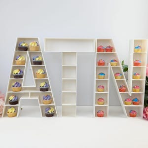 May include: White cupcake stand shaped like the letters "ATN". Each letter has multiple shelves to hold cupcakes. The cupcakes are decorated with different coloured frosting.