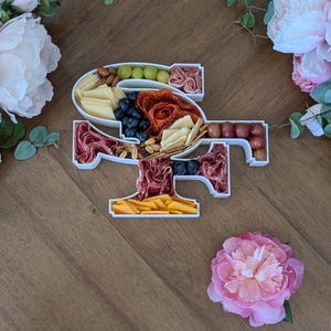 Peut inclure: Un plateau de service en céramique blanche en forme de chiffre "49" rempli de diverses collations, notamment du fromage, du salami, des raisins et des craquelins. Le plateau est posé sur une surface en bois avec des fleurs roses de chaque côté.