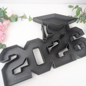 May include: A dark gray graduation cap and the number "2023" cookie cutter. The cutter is made of plastic and is on a white surface. Pink and white flowers are in the background.
