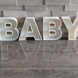 May include: White plastic letters spelling out the word "BABY" on a gray tiled floor.