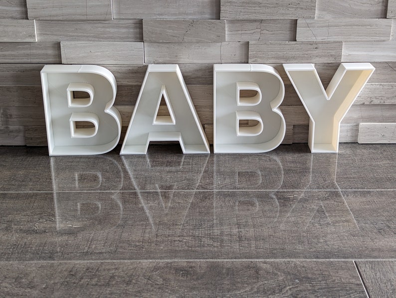 May include: White plastic letters spelling out the word "BABY" on a gray tiled floor.