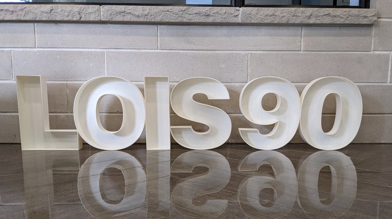May include: White, freestanding letters spelling "LOIS 90" on a tiled floor. The letters are hollow and have a 3D effect.
