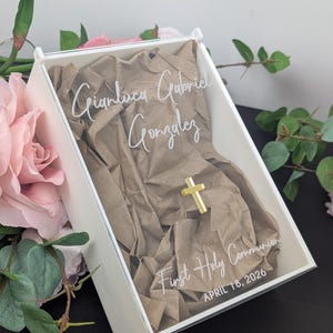 May include: A white box with a clear lid, containing brown paper and a gold cross. The lid has the text "Gianluca Gabriel Gonzalez" and "First Holy Communion APRIL 18, 2026" in white script. Pink and green floral accents surround the box.