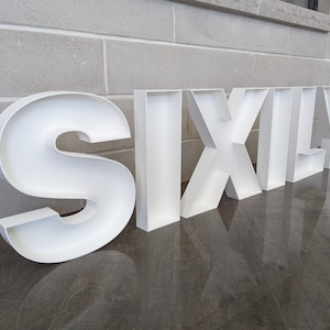 May include: White, hollow, plastic letters spelling out the word "SIXLY" on a gray tiled floor.