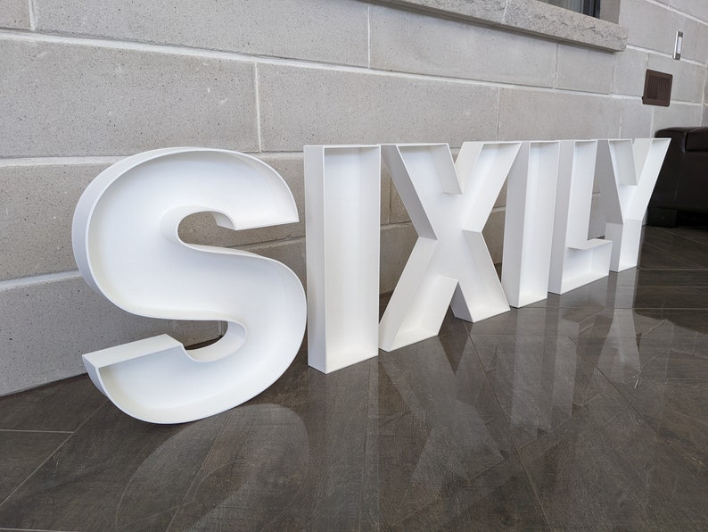 May include: White, hollow, plastic letters spelling out the word "SIXLY" on a gray tiled floor.