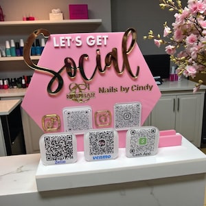 May include: A pink hexagonal sign with the words "LET'S GET Social" in white and gold lettering. Below are QR codes for social media and payment apps. The sign is for "Nails by Cindy".
