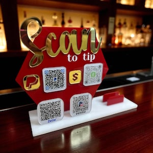 May include: A red acrylic sign with gold lettering that says "Scan to tip." The sign has several QR codes for payment apps like PayPal, Zelle, and Venmo. The sign is on a white base.