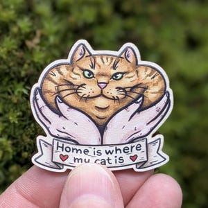 May include: A wooden pin featuring an illustration of a round orange tabby cat held by two hands. Below the cat, a banner displays the text "Home is where my cat is" with a small red heart on each side of the word "my" in the phrase.