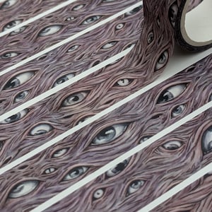 May include: Washi tape with a repeating pattern of detailed eyes in brown and grey. The tape is rolled and unrolled, with white stripes separating the eye-covered sections. The design has a surreal, artistic quality. The tape is suitable for crafts.