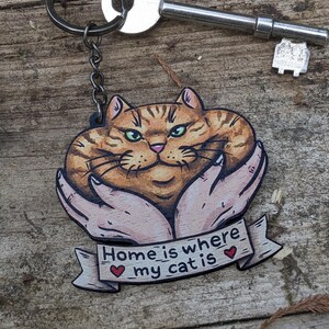 May include: A cat-themed keychain featuring a tabby cat design held in hands, with the text "Home is where my cat is." The keychain is attached to a metal ring and chain, with a key nearby.