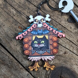 May include: A whimsical Halloween-themed keychain featuring a black cat in a house-shaped design. The house has a skull, bat, and decorative elements. The keychain is attached to a metal chain and key. The color palette includes brown, pink, and yellow.