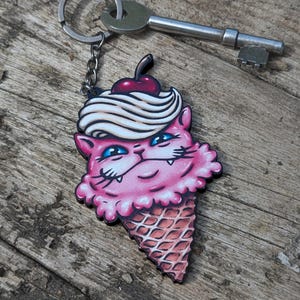 May include: A pink cat-shaped keychain with blue eyes, a cherry on top, and a white swirl design, resembling ice cream. The keychain is attached to a silver chain and key. The cat is in a waffle cone.