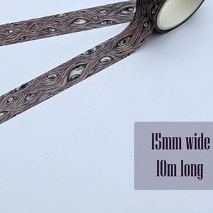 K&ouml;nnte beinhalten: Dekoratives Washi-Tape mit einem sich wiederholenden Muster aus offenen Augen auf braunem Hintergrund. Das Klebeband ist 15 mm breit und 10 m lang, wie auf dem Etikett angegeben. Ein Teil des Bandes ist abgerollt.