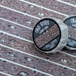 K&ouml;nnte beinhalten: Zwei Rollen Washi-Tape mit dem Text "OLD GODS WASHI TAPE" und einem Design aus mehreren Augen. Der Hintergrund zeigt ein sich wiederholendes Muster aus Augen und wei&szlig;en Streifen. Das Klebeband ist in Papier verpackt.
