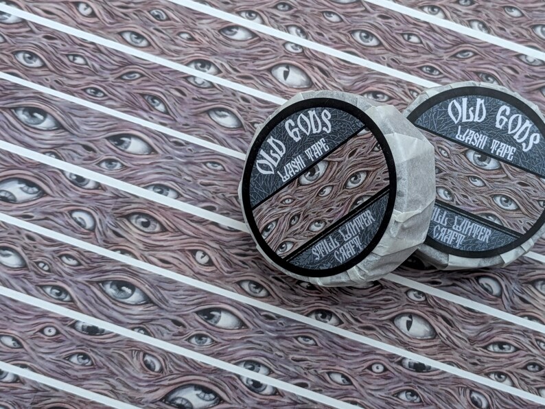 K&ouml;nnte beinhalten: Zwei Rollen Washi-Tape mit dem Text "OLD GODS WASHI TAPE" und einem Design aus mehreren Augen. Der Hintergrund zeigt ein sich wiederholendes Muster aus Augen und wei&szlig;en Streifen. Das Klebeband ist in Papier verpackt.